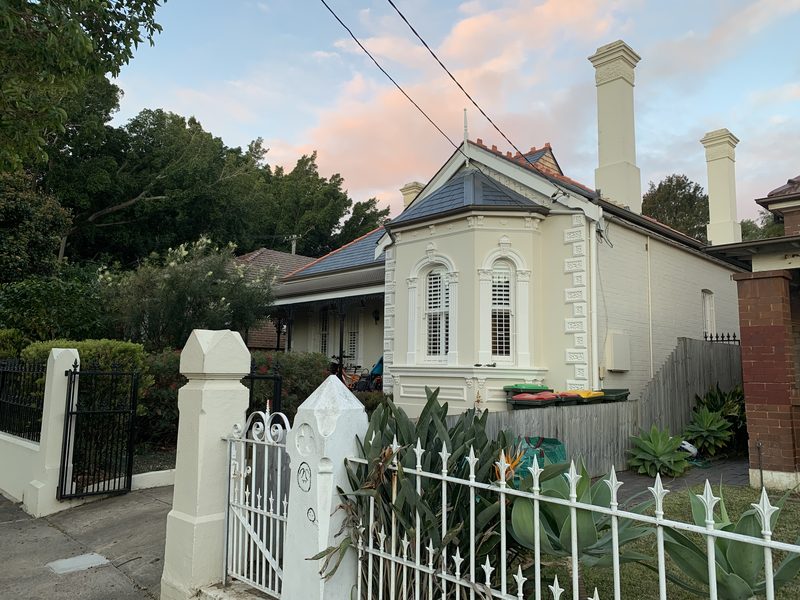A Victorian cottage behind its white fence and gate, with a bay window and tall chimneys