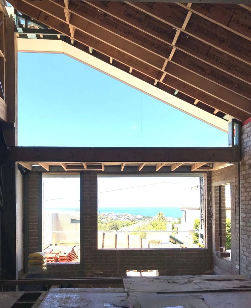 An extension under construction: a new timber roof frame above brick openings that frame a view of the ocean