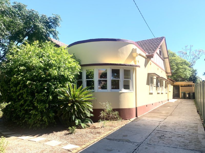A rendered inter-war house with a curved bay window and a tiled roof, behind a leafy garden